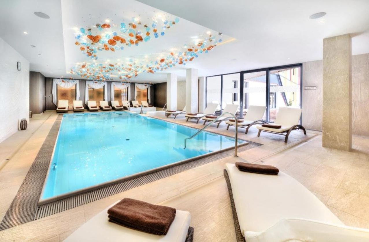 A swimming pool with lounge chairs, a ceiling with decorative elements, and large windows.