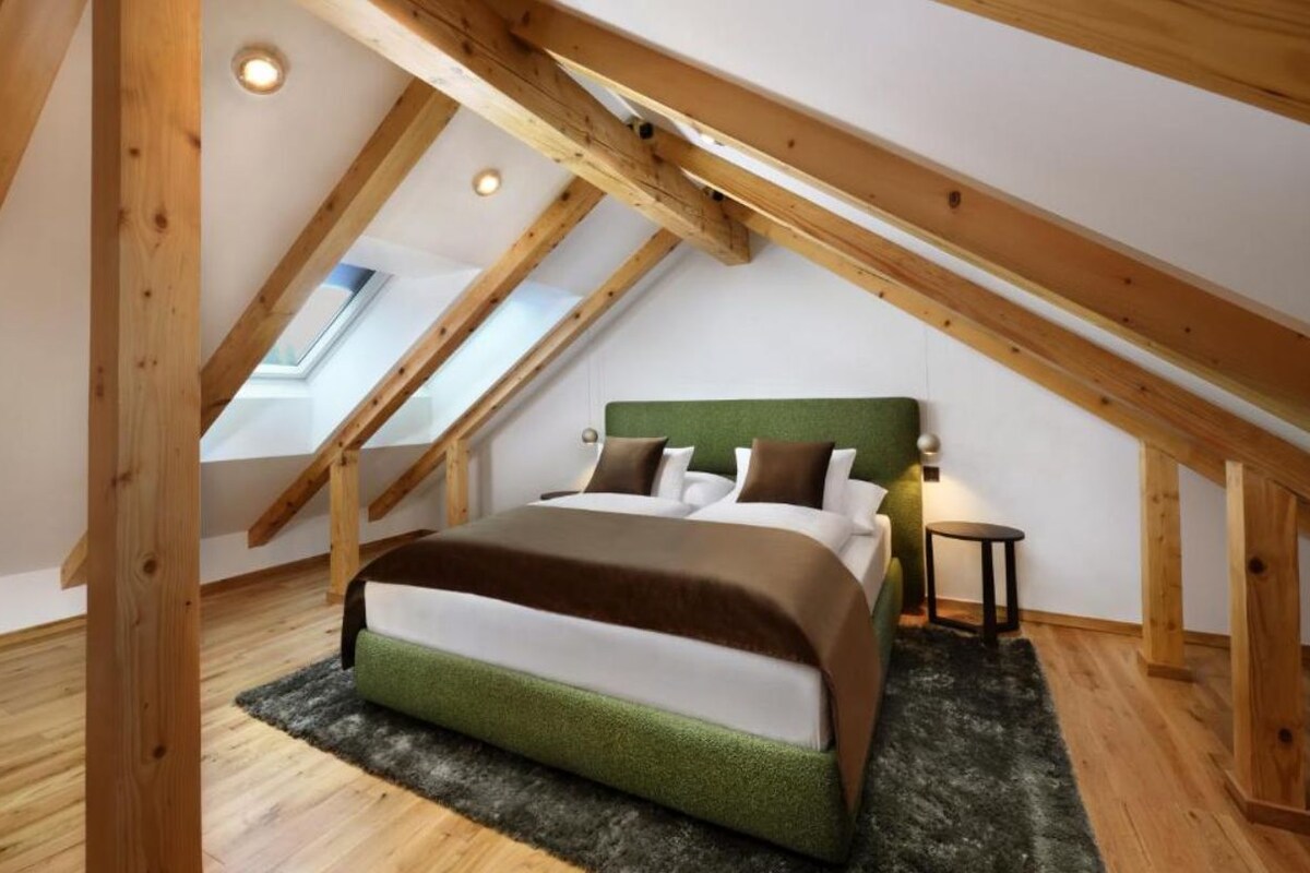 Ein Schlafzimmer mit schrägen Wänden, Holzbalken, einem Bett mit Kissen, einem kleinen Tisch und einem Teppich.