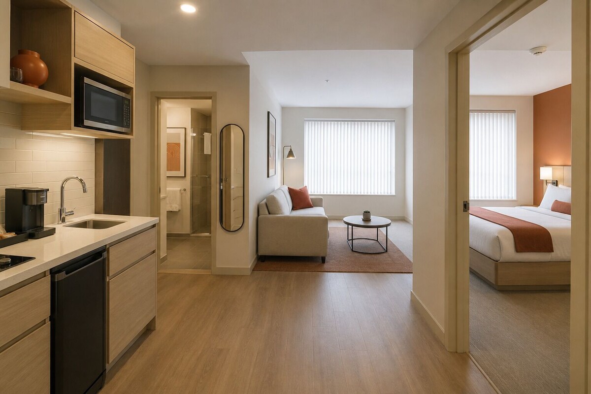 Een moderne hotelkamer met een kitchenette, een bank, een klein rond tafeltje en een bed met een bruine plaid.