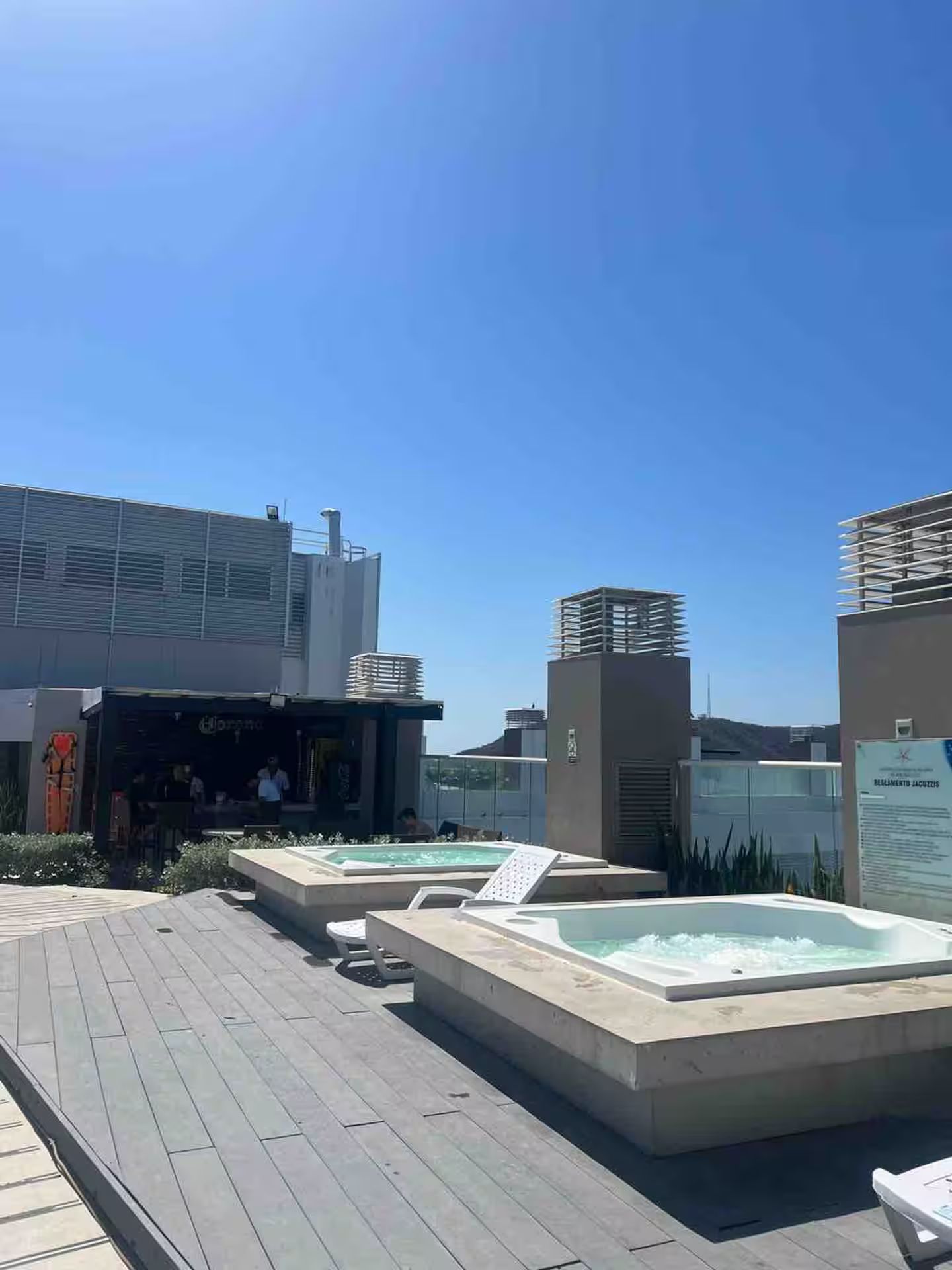 A rooftop with two hot tubs, a wooden deck, and a modern building with a sign.
