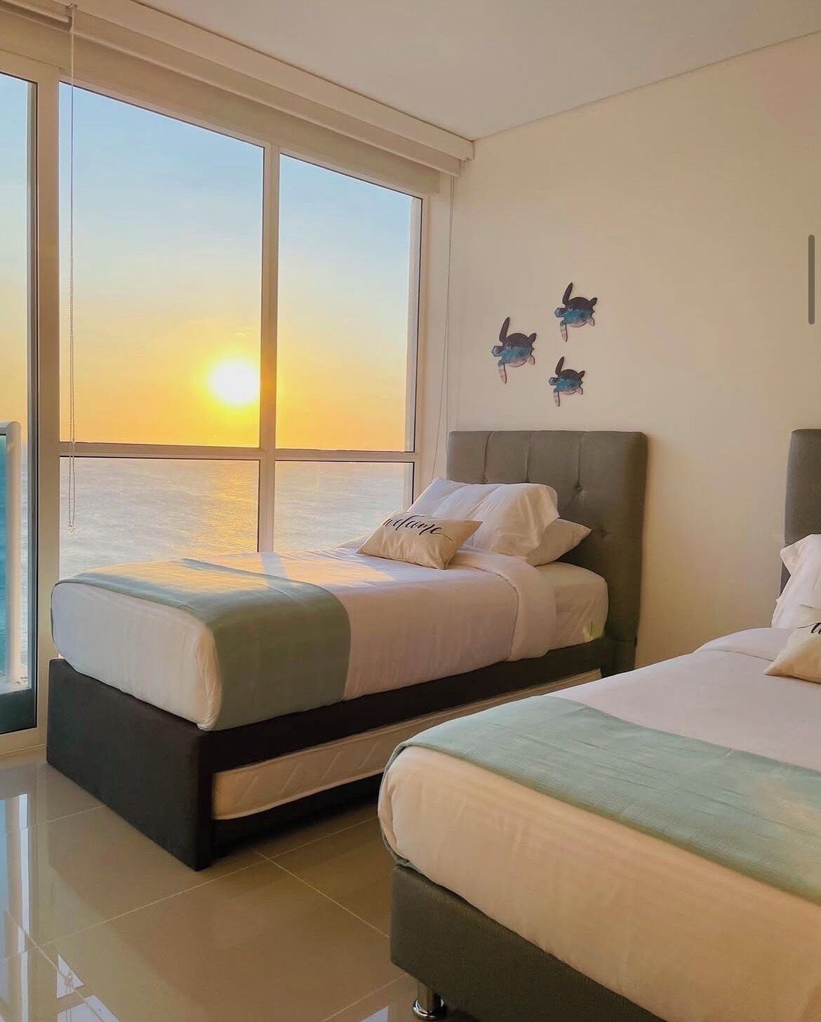 Ein Hotelzimmer mit zwei Betten, einem großen Fenster mit Blick auf den Sonnenuntergang und Wanddekorationen mit Schildkrötenmotiven.