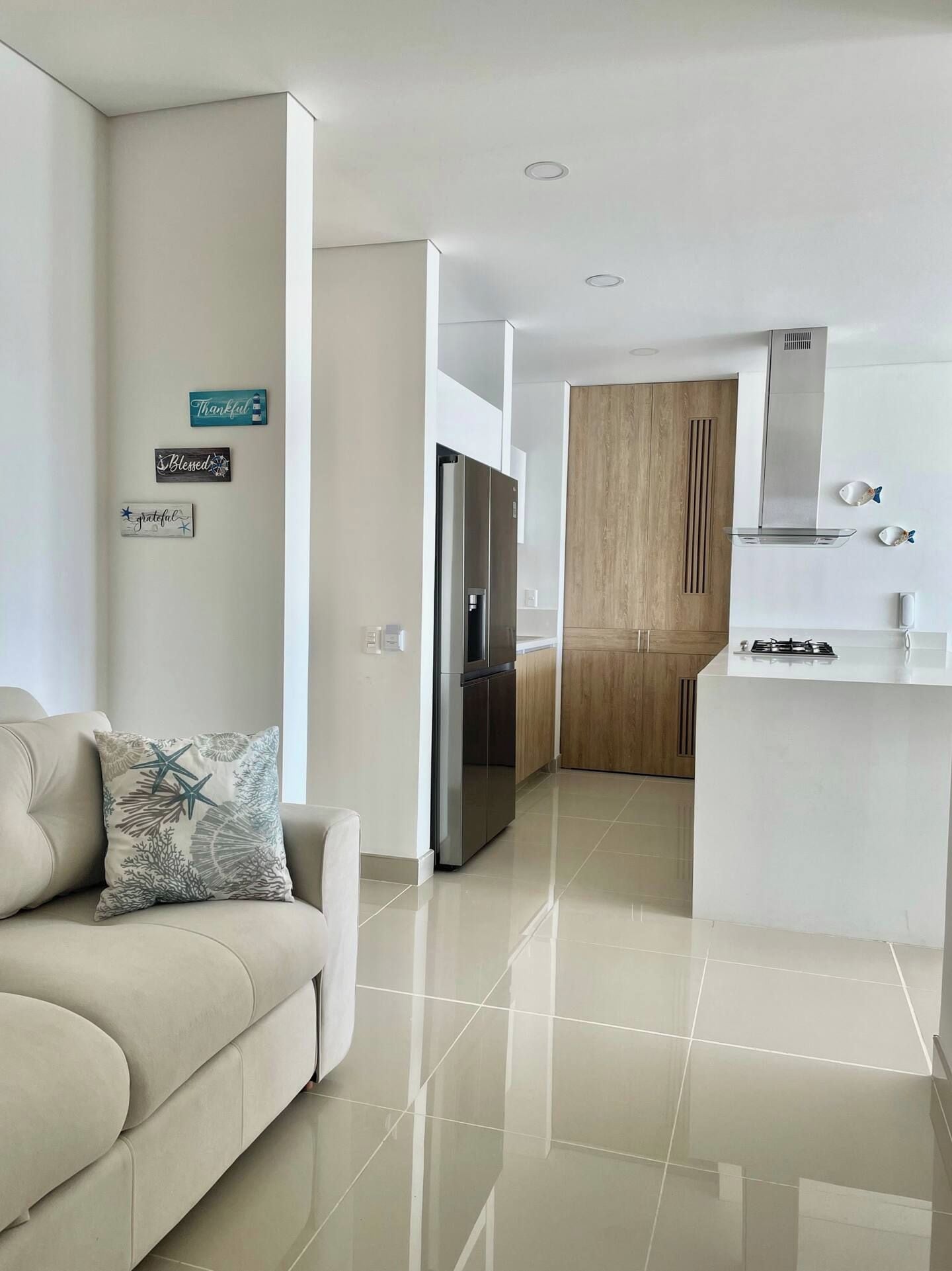 Sala moderna com sofá e almofadas decorativas, conectada a uma cozinha com armários de madeira e fogão a gás.