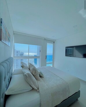 Ein modernes Schlafzimmer mit einem großen Bett, einer Couch, einem an der Wand befestigten Fernseher und Meerblick durch große Fenster.