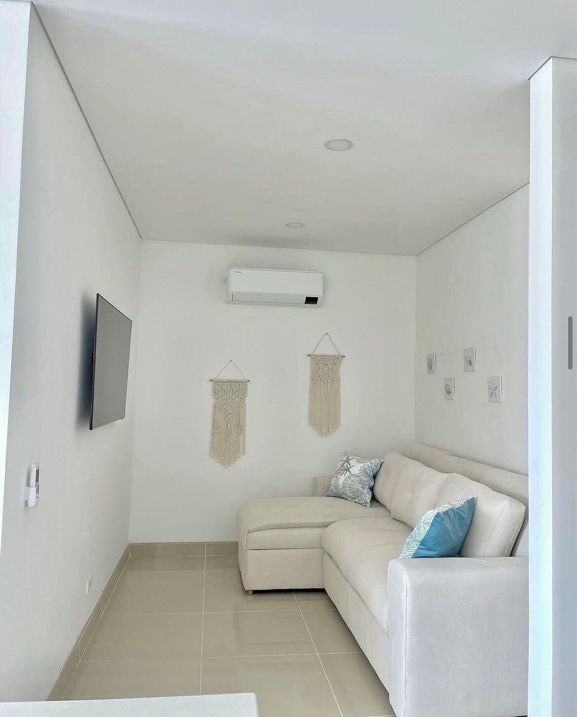 A modern living room with a beige sectional sofa, a flat-screen TV mounted on the wall, and an air conditioning unit.