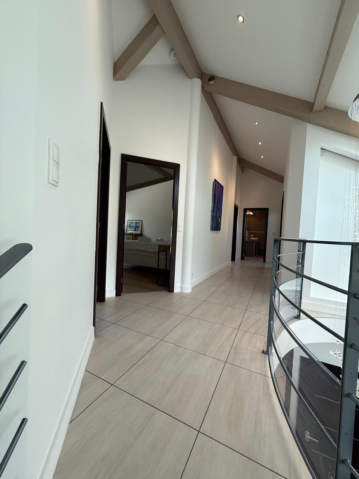 A spacious hallway with a high ceiling, a glass railing on the right, and a door leading to a room with a bed.