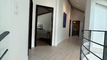 A spacious hallway with a high ceiling, a glass railing on the right, and a door leading to a room with a bed.