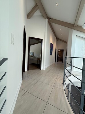 A spacious hallway with a high ceiling, a glass railing on the right, and a door leading to a room with a bed.