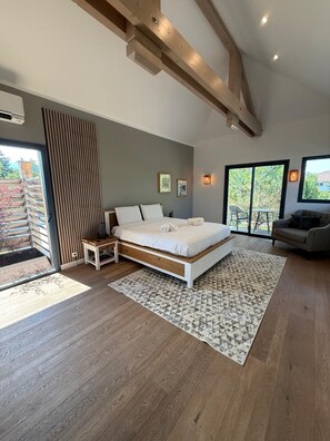 A modern bedroom with a large bed, a wooden floor, a view through a sliding door, and a sofa in the corner.