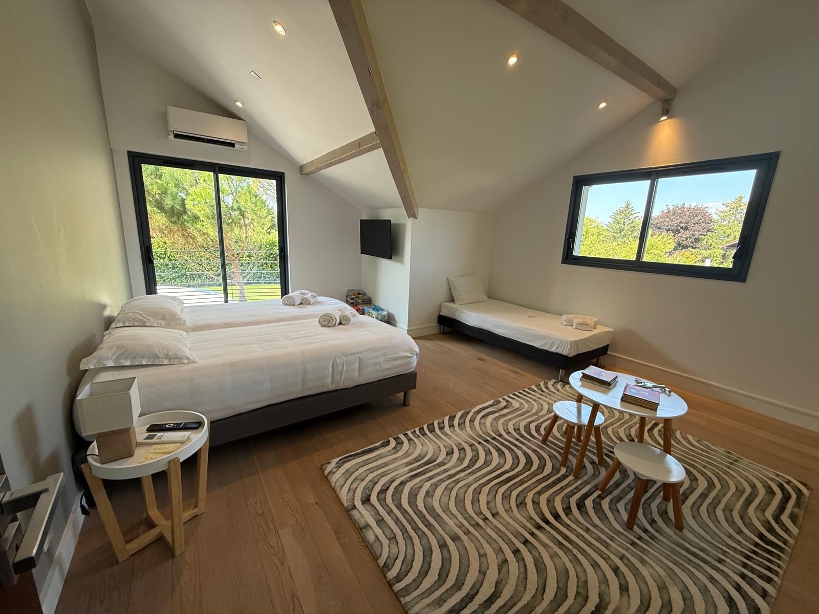 A bedroom with a large bed, a smaller bed, a desk with chairs, a TV, and large windows with a view of greenery.