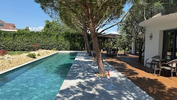 Une piscine avec une terrasse en bois, un espace repas aménagé avec du mobilier d’extérieur et un grand arbre qui offre de l’ombre.