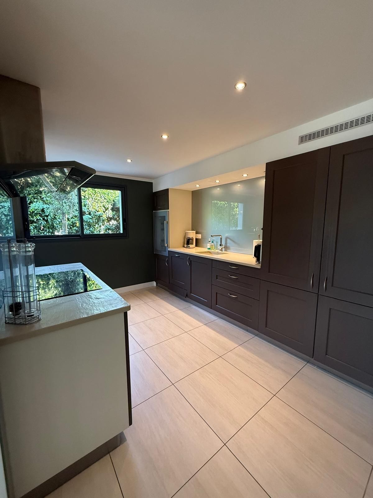 A modern kitchen with dark cabinets, a central island, and a view of greenery outside.