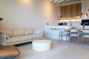 Ein modernes Wohnzimmer mit Sofa, Hocker und einem Essbereich mit Stühlen, im Hintergrund eine Küche.