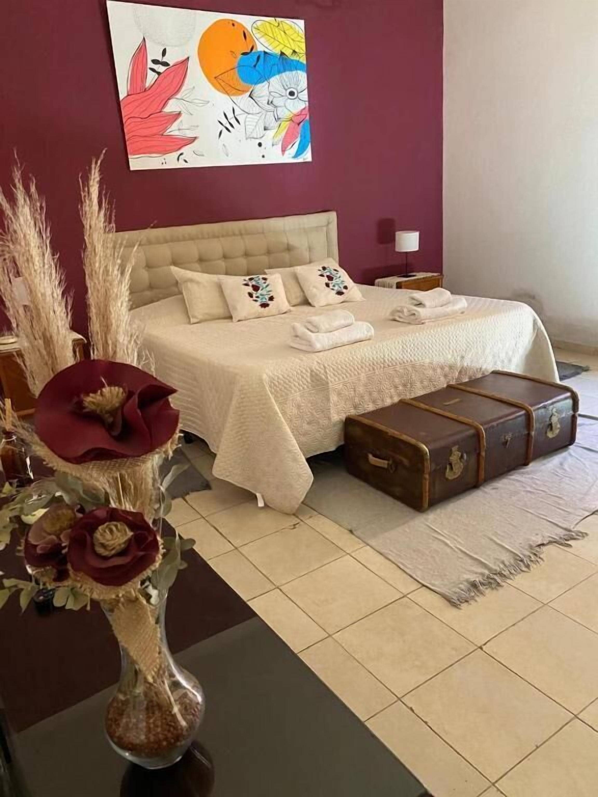 Un dormitorio con una cama, una mesita de noche, un jarrón con flores y una maleta.