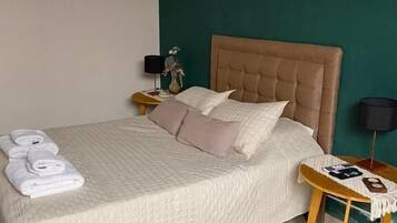 A neatly made bed with a beige quilt, a bedside table with a lamp, and a wall with a colorful abstract painting.