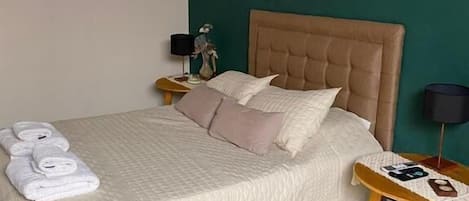 A neatly made bed with a beige quilt, a bedside table with a lamp, and a wall with a colorful abstract painting.