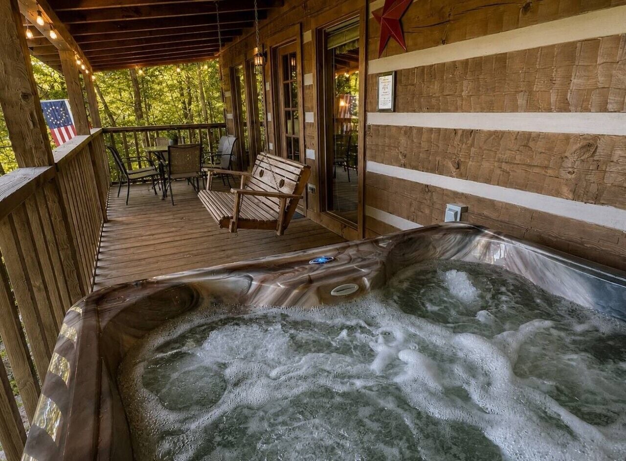 A hot tub on a wooden deck with chairs and a table, surrounded by a wooden fence and trees.