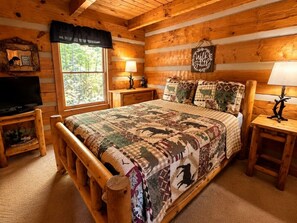 A log cabin bedroom with a wooden bed, two bedside tables, a television, and a window with a view of trees. - Mtn Views + Hot Tub | 10 Min to Pigeon Forge (Sevierville)