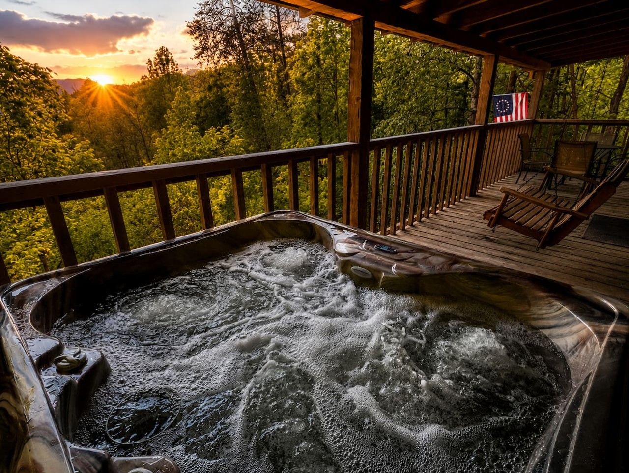 Een hottub met een houten terras en stoelen, omringd door bomen en uitzicht op een zonsondergang.