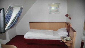 Una camera d'albergo piccola con un letto singolo, un comodino con il telefono, una finestra con tende e un quadro appeso al muro.