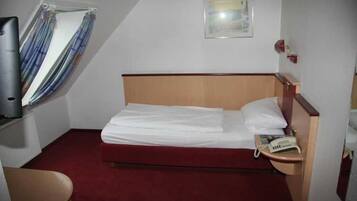 Een kleine hotelkamer met een enkel bed, een nachtkastje met telefoon, een raam met gordijnen en een aan de muur hangend schilderij.