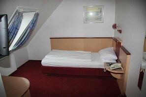 Quarto de hotel pequeno com uma cama de solteiro, mesa de cabeceira com telefone, janela com cortinas e um quadro pendurado na parede.