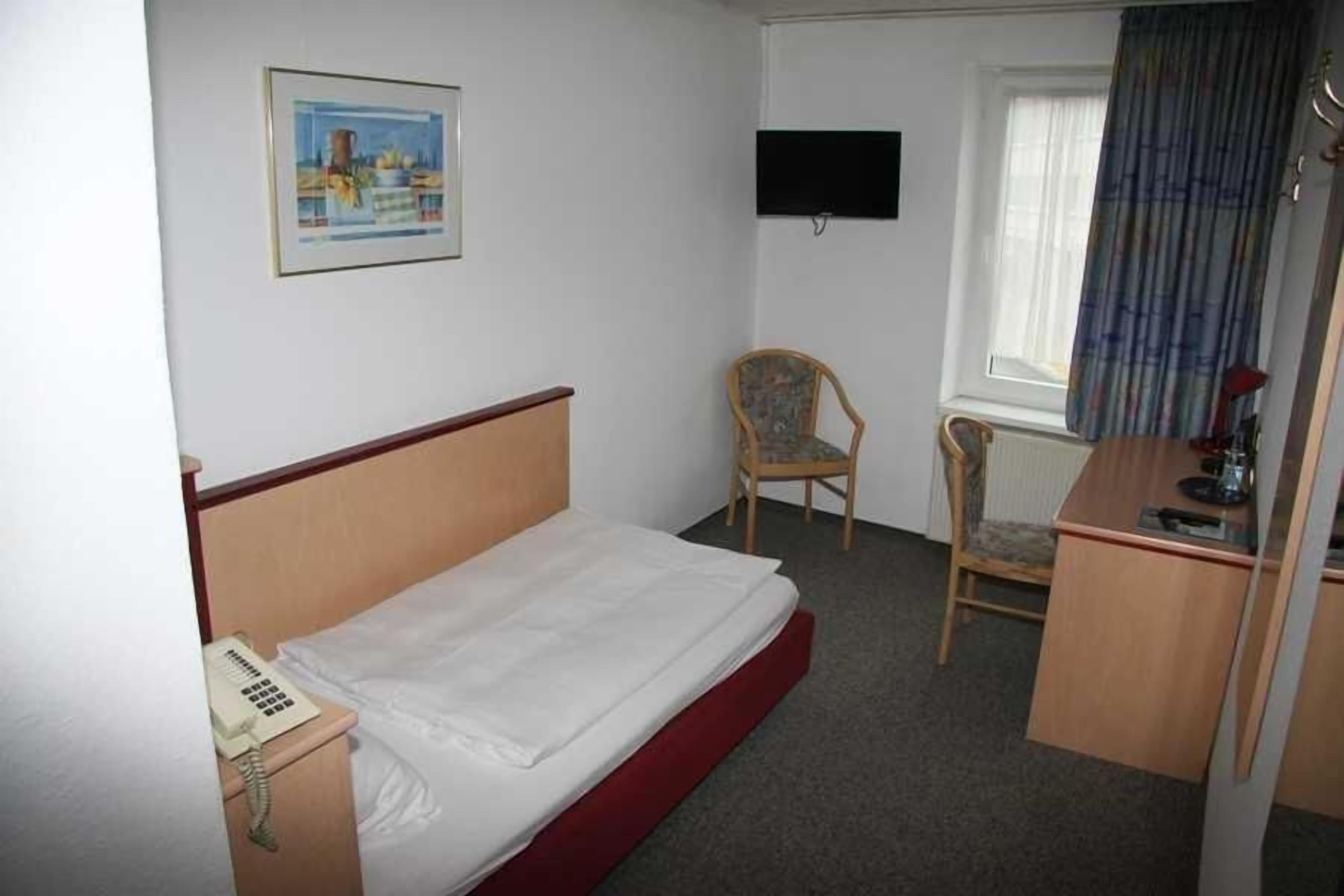 Una habitación de hotel con cama, escritorio, silla, teléfono, televisor montado en la pared y ventana con cortinas.