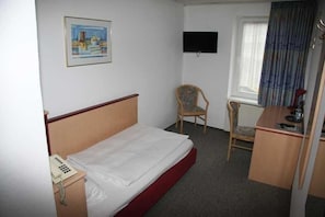 Ett hotellrum med en säng, ett skrivbord, en stol, en telefon, en tv som är monterad på väggen och ett fönster med gardiner.