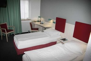 Quarto de hotel com duas camas, uma escrivaninha, uma cadeira e uma janela com cortinas.