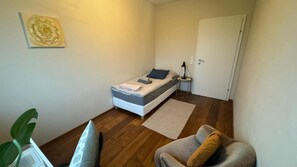 Ein kleines Zimmer mit einem Einzelbett, einem Nachttisch, einem Stuhl, einer Pflanze und einer Tür.
