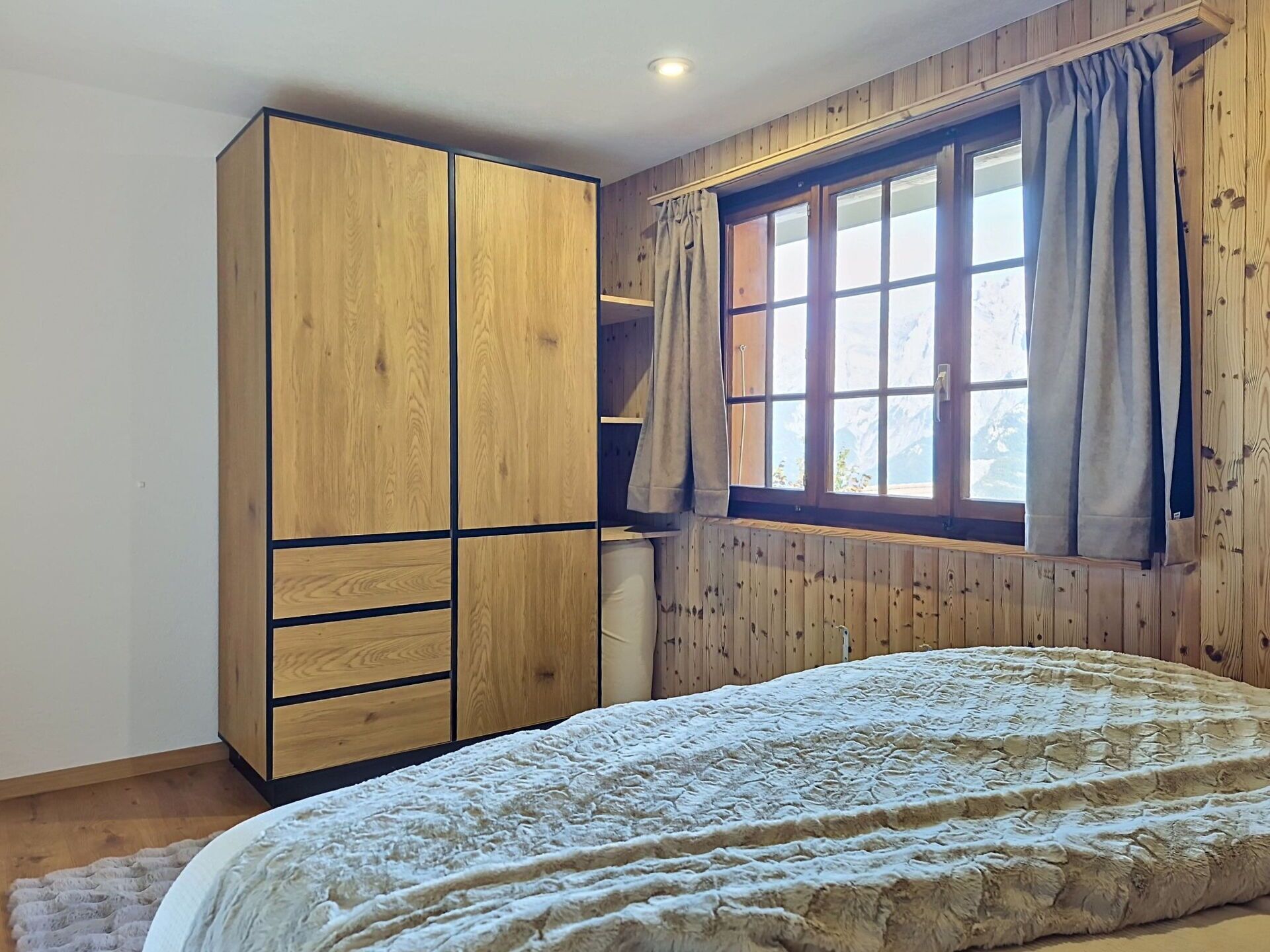 A bedroom with a large wardrobe, a bed with a textured blanket, a window with curtains, and wooden paneling on the walls.