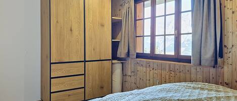 A bedroom with a large wardrobe, a bed with a textured blanket, a window with curtains, and wooden paneling on the walls.