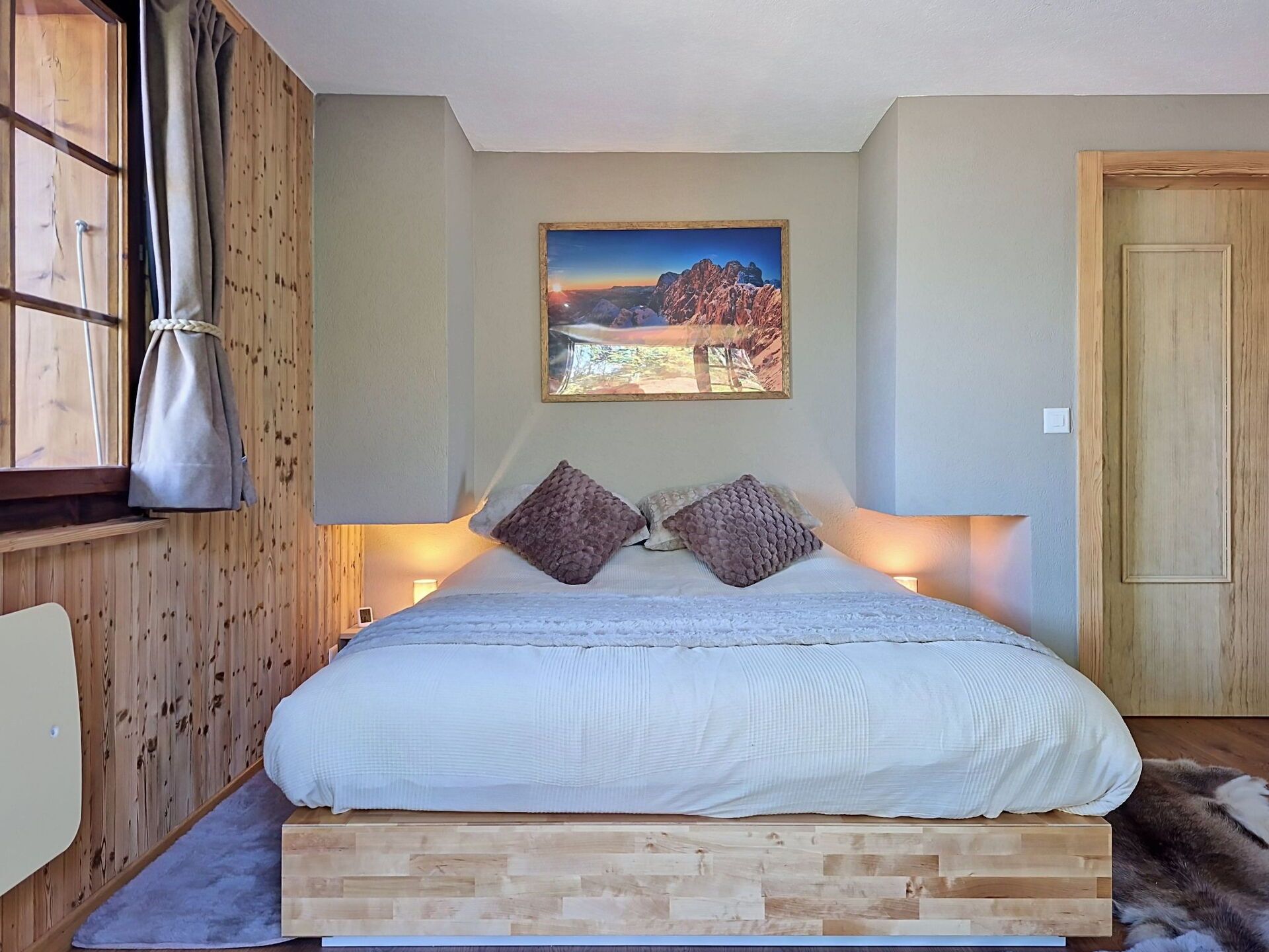 Ein Schlafzimmer mit hölzernem Kopfteil, einem Bett mit weißer Bettwäsche, zwei strukturierten Kissen, einem Landschaftsbild und einem Fenster mit Vorhängen.