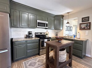 Una cocina con armarios verde oscuro, un refrigerador de acero inoxidable, un microondas, una estufa y una mesa de madera con una cesta tejida.