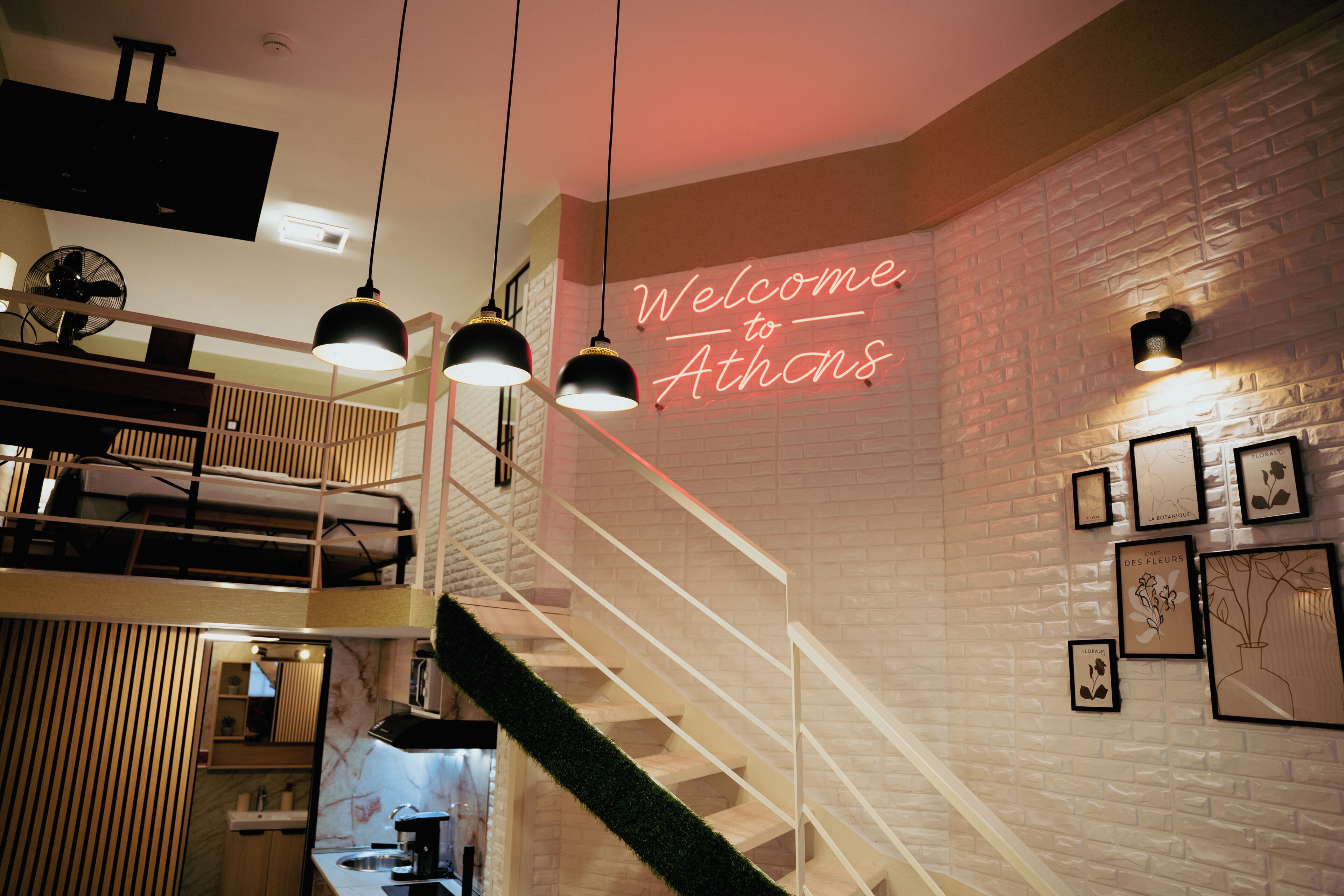 A modern interior with a staircase, a neon sign reading 'Welcome to Athens,' and framed botanical artwork on the wall.