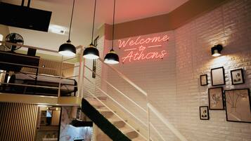 A modern interior with a staircase, a neon sign reading 'Welcome to Athens,' and framed botanical artwork on the wall.