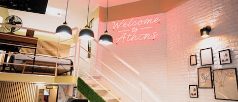 A modern interior with a staircase, a neon sign reading 'Welcome to Athens,' and framed botanical artwork on the walls.