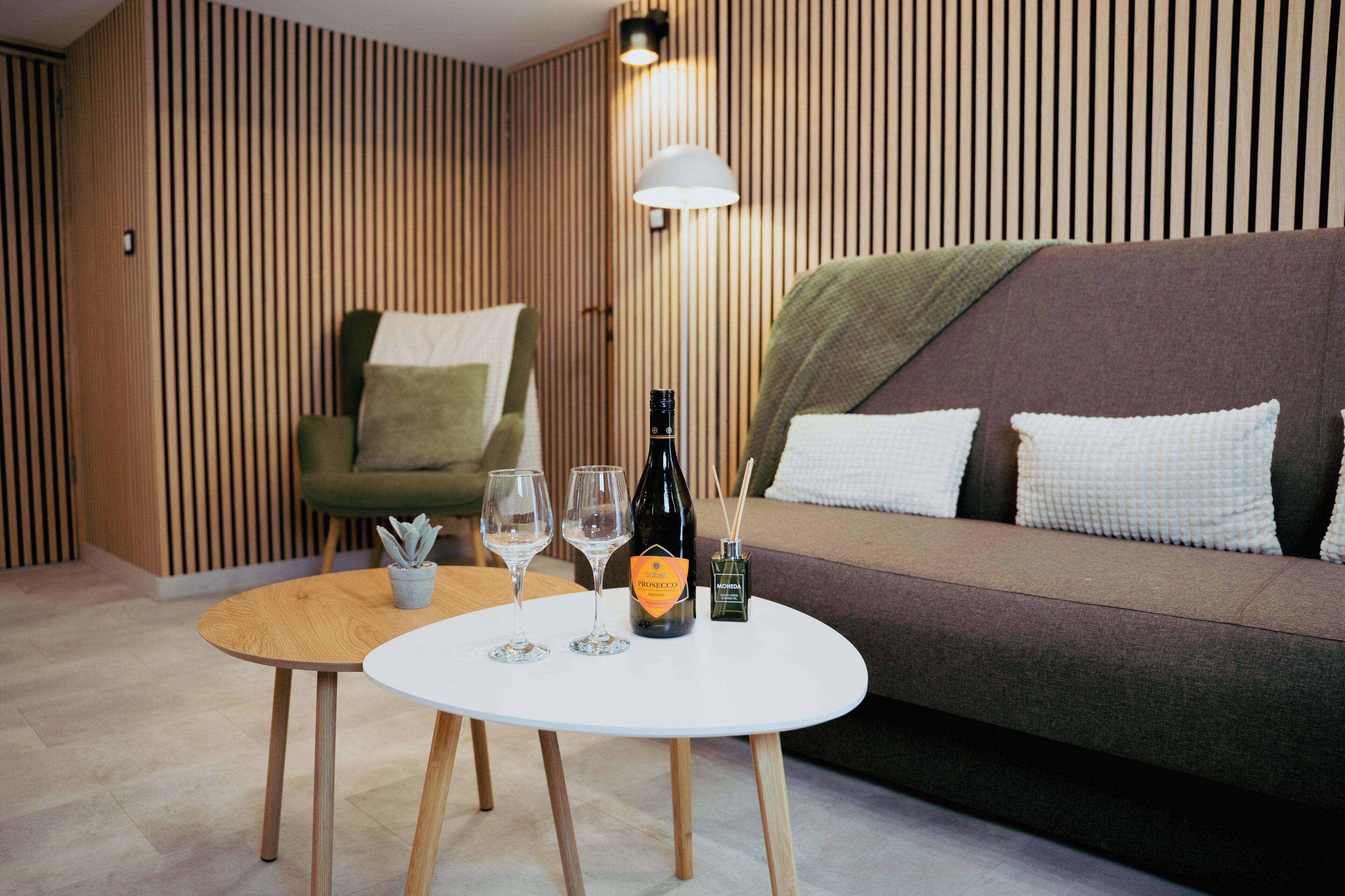 Moderni huone, jossa on pyöreä puinen pöytä, kaksi viinilasia, pullo Prosecco-kuohuviiniä ja pieni ruukkukasvi.