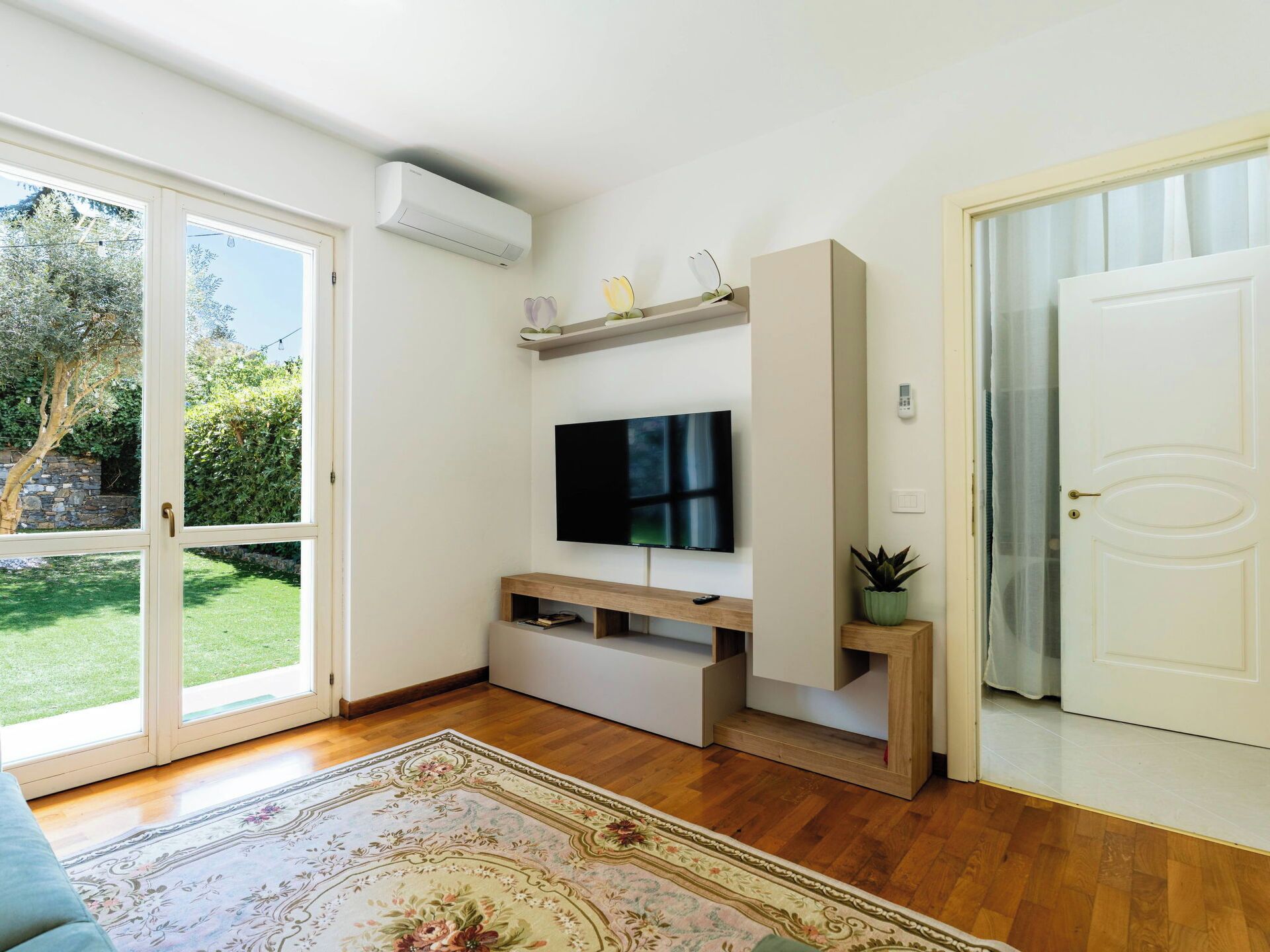 Un salon moderne doté d’un téléviseur à écran plat, d’un meuble de rangement en bois et d’une grande fenêtre donnant sur un paysage verdoyant.