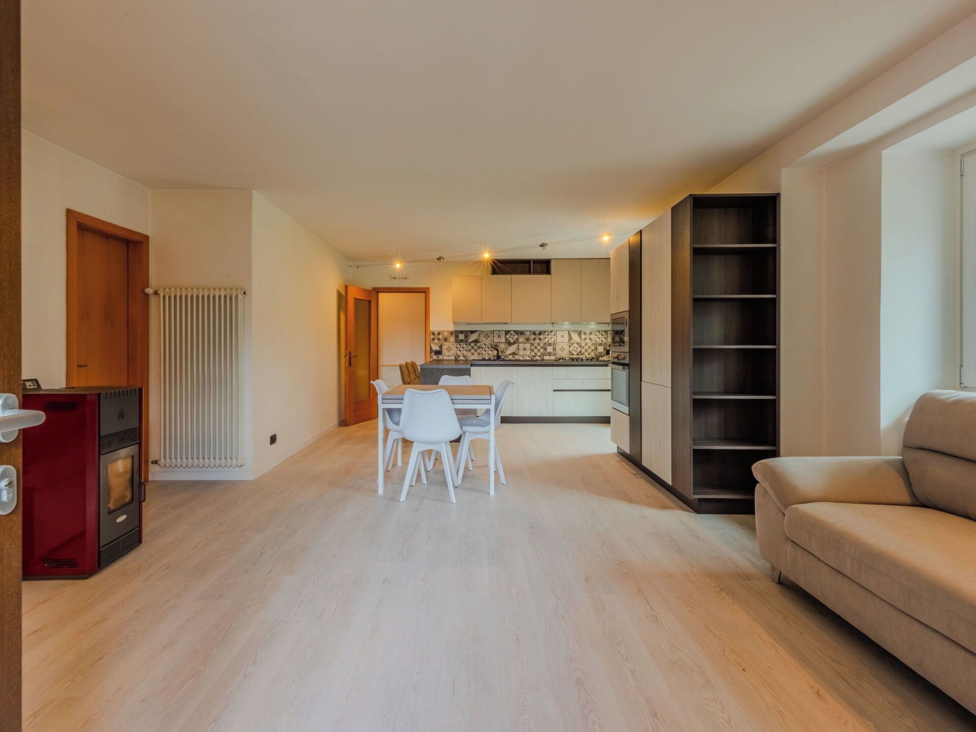 A modern living room with a sofa, a dining area with a table and chairs, and a kitchen with a tiled backsplash.