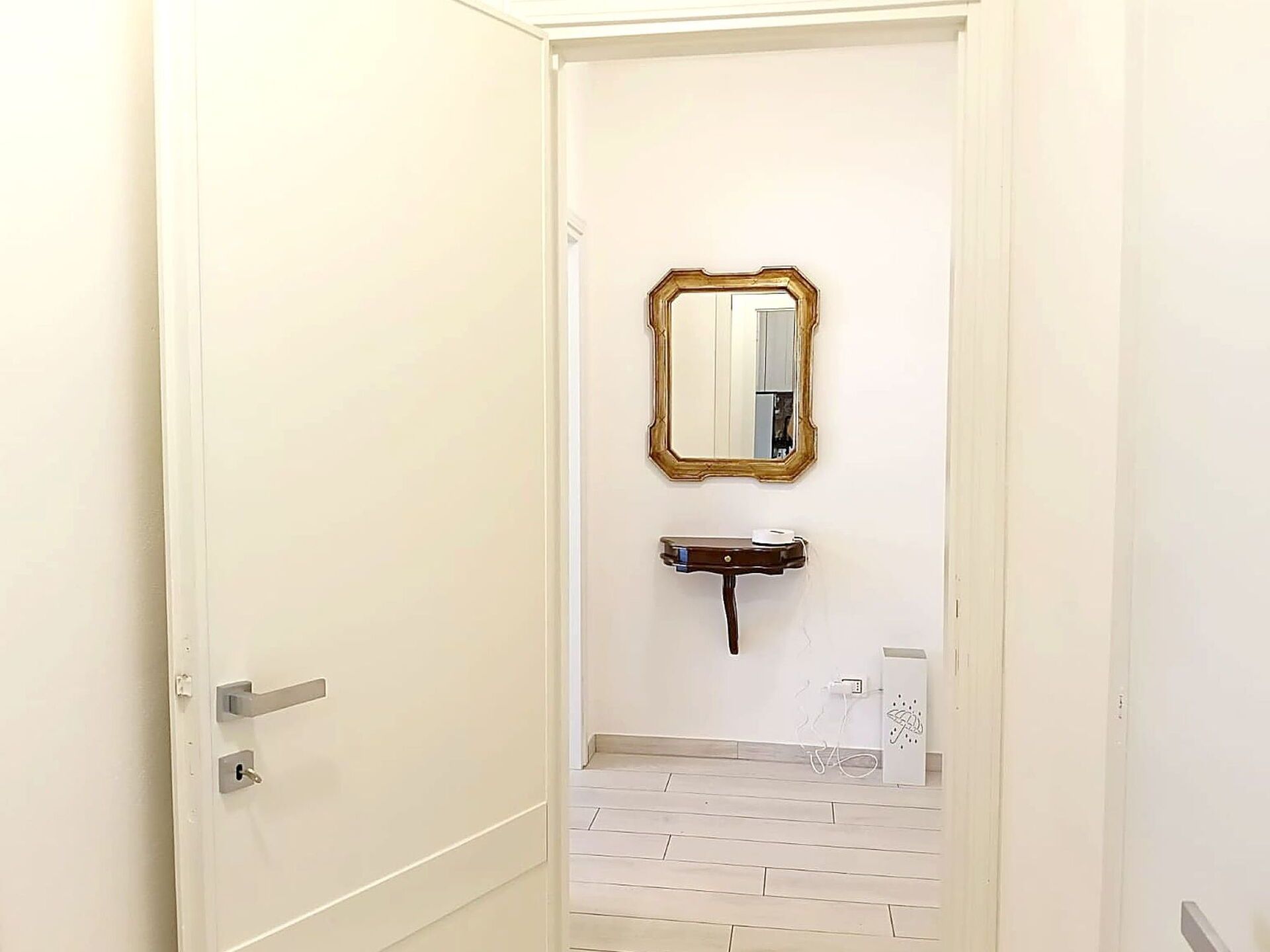 Un couloir avec une porte blanche, un miroir, une petite étagère en bois et un sol carrelé de carreaux blancs.