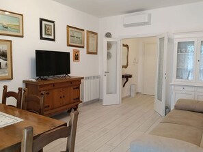 Een woonkamer met een televisie, een houten kast, een eettafel met stoelen en een bank. Er hangen ingelijste foto's aan de muur en een deur die naar een andere kamer leidt.