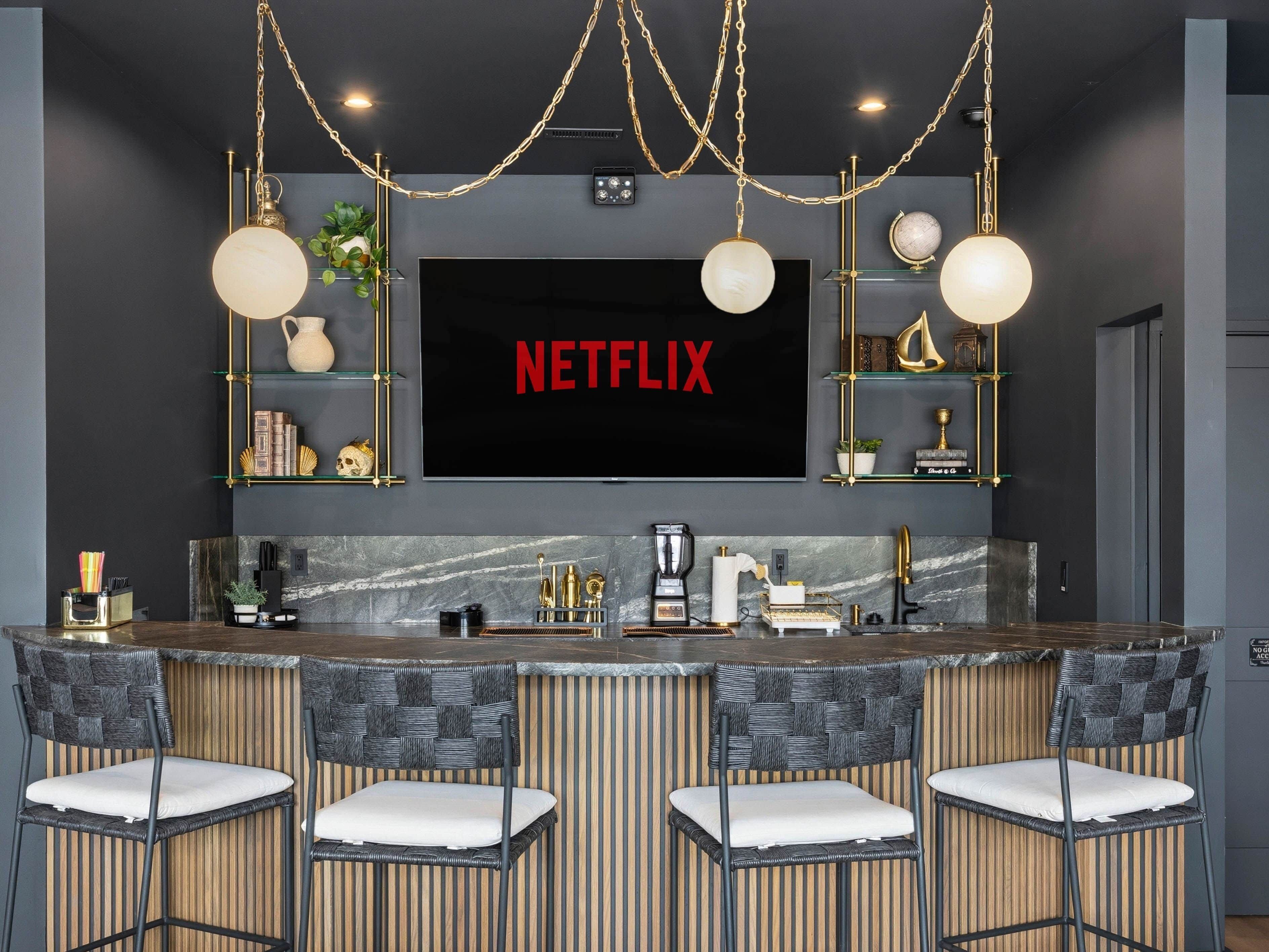 Une cuisine moderne dotée d’un grand téléviseur à écran plat affichant le logo « NETFLIX », de tabourets de bar et d’objets décoratifs disposés sur des étagères.