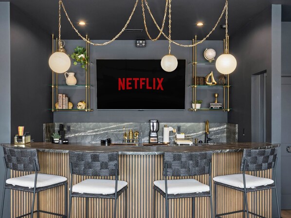 Cocina moderna con una televisión de pantalla plana grande mostrando 'NETFLIX', taburetes de bar y objetos decorativos en estantes.