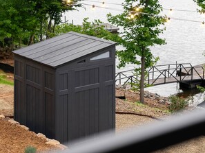 Una caseta oscura con techo inclinado, ubicada cerca del agua, con un muelle y un puente al fondo.