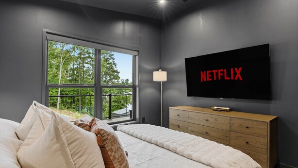 A bedroom with a large window, a bed with pillows, a wooden dresser, and a TV displaying the Netflix logo. - The Black Pearl - Lake Bowen - Luxury Waterfront (Inman)