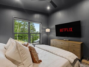 Ein Schlafzimmer mit großem Fenster, einem Bett mit Kissen, einer Holzkommode und einem Fernseher, der das Netflix-Logo zeigt.