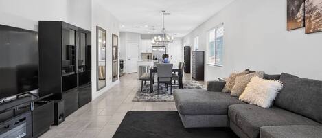 A modern living room with a large flat-screen TV, a grey sofa, and a dining area with a chandelier.