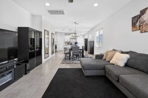 A modern living room with a large flat-screen TV, a grey sofa, and a dining area with a chandelier. - Mins to Banner* St. Mary’s* El Rio* TUS Airport (Tucson)