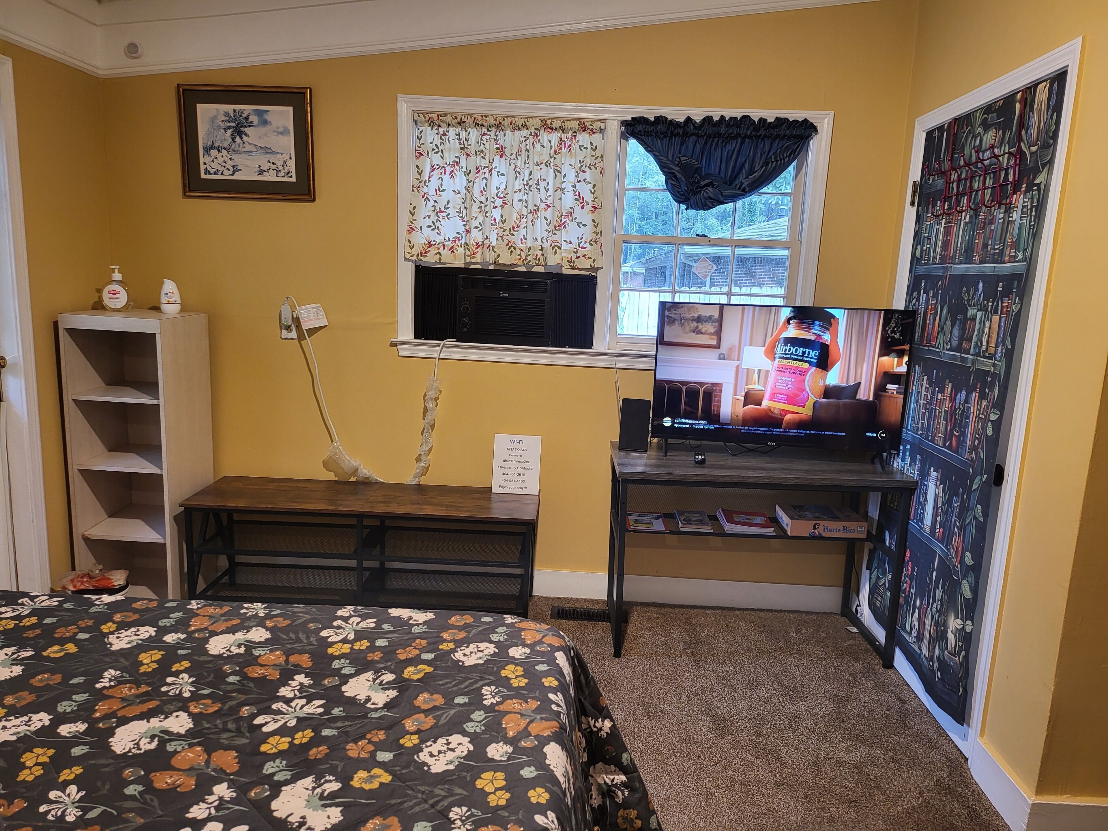 A bedroom with a bed, a desk with a computer, a window with curtains, and a bookshelf.