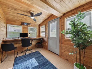 A cabin interior with a wooden ceiling, a ceiling fan, a desk with a computer, two chairs, a door, and a potted plant.